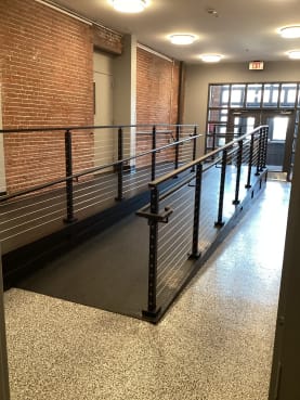 Black posts with cable infill and grab rails line an interior ramp in a renovated brick building.