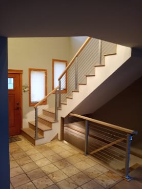 Cable railing with a wood handrail climbs a white stair and guards the opening beneath it beside a wood entry door.