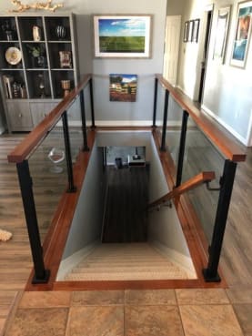 Black posts with glass panels and a wood cap rail enclosing a stairwell opening on a tiled floor.