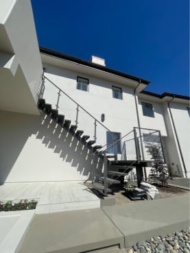 An exterior mono stair with dark treads and glass railing climbs a white stucco facade.