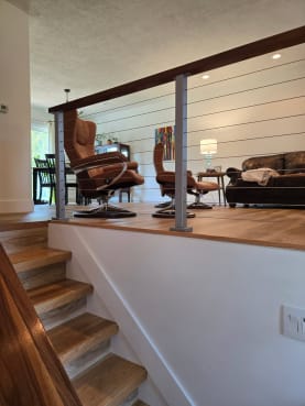 Silver-post cable railing with a wood cap rail edges a raised living area above an oak staircase.