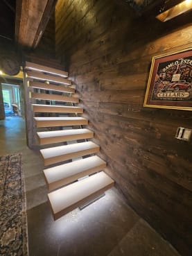 Light wood floating treads with LED undersides climb a dark reclaimed-wood wall in a stone-floored lower level.