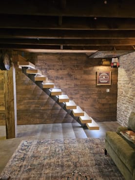 Light wood floating treads on a mono stringer rise along a reclaimed barnwood wall in a cozy basement lounge.