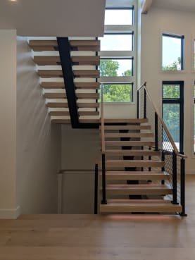 White oak treads on a black mono stringer turn at a landing with cable railing and wood handrail, framed by tall windows.