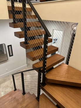 Looking down wood floating winder treads beside black-post cable railing in a bright entry.