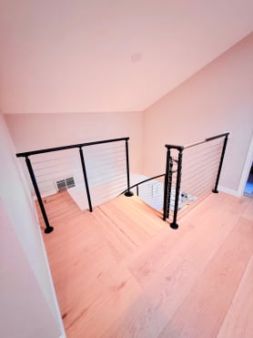 Black posts with cable infill guard a stair opening on light hardwood floors.