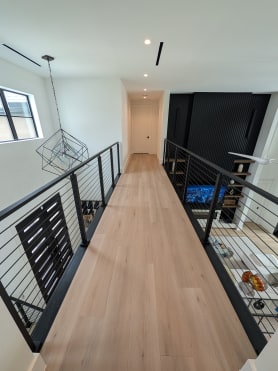 Upper floor oak bridge walkway lined on both sides by black cable railing above an open living area.