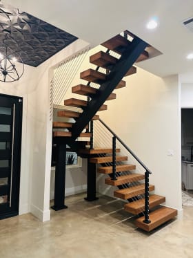 Warm wood floating treads on a black mono stringer switch back at a landing, with black cable railing beneath a decorative ceiling.