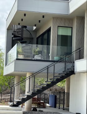 Elevated modern home with a black exterior stair with cable railing, glass balcony railing and spiral stair above.