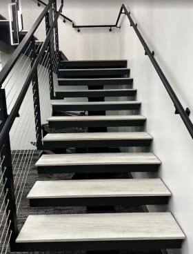 Gray treads on a black mono stringer between black cable railing and a wall-mounted black handrail in an office stairwell.