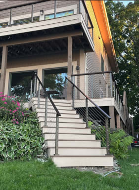 Bronze-post cable railing lines wide composite exterior stairs leading up to a deck on a lake-style home.
