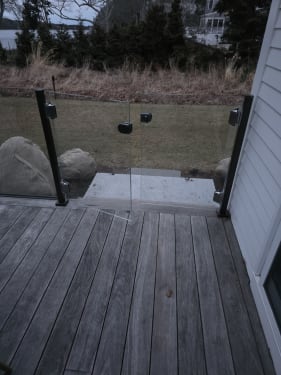 Frameless glass double gate between black posts opens from a weathered wood deck to a stone path.