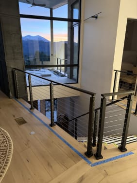 Black-post cable railing wraps a stair opening in front of tall windows framing a mountain sunset