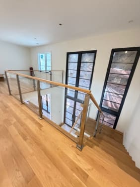 Brushed stainless posts and glass panels topped with an oak handrail guard a stair opening in a bright hallway with black steel windows.