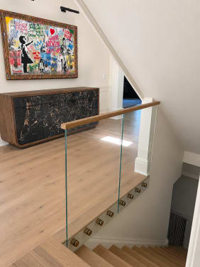 Frameless glass panels with brass standoff pins and an oak handrail line a white-oak stair in a paneled white entry.
