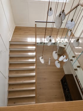 View from above of a white-oak stair with a frameless glass guard on brass standoff pins beneath a cascading disc chandelier.