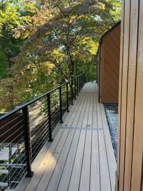 Black cable railing runs along a narrow composite deck walkway beside a wood-sided home among trees.