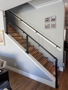 Black cable railing mounted to a white knee wall alongside a stained wood stair with wall handrail.