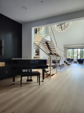 Oak floating stair with frameless glass guard and wood handrail seen past a black grand piano in a bright living room.