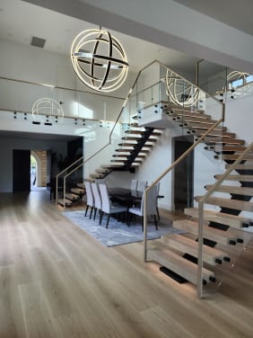 Pair of oak floating stairs on black mono stringers with frameless glass, wood handrails and LED glow framing a dining room under orb chandeliers.
