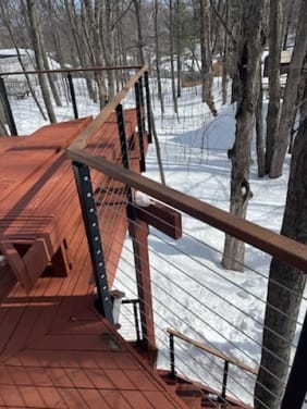 Slim side mount cable railing with Ipe handrail follows a red deck and stair through snowy trees.