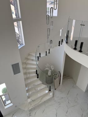 Curved glass panels on black talon spigots follow a sweeping white marble stair beneath a crystal chandelier.