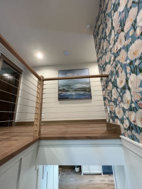 Oak newels and cap rail with cable infill around a stair opening between white wainscoting and floral wallpaper.