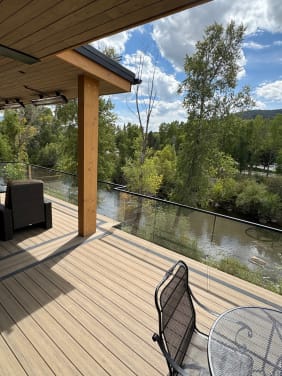 Frameless glass on black standoff pins edges a covered composite deck above a tree-lined river.