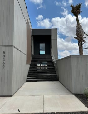 Wide black steel open-riser stair rises between gray-clad walls toward an upper entry under blue sky.