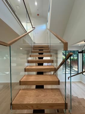 Looking up a floating white oak stair on a black mono stringer with glass panels and wood cap handrails on both sides.