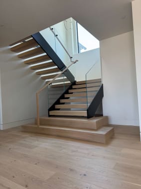 Light oak floating treads on a black mono stringer turn at a landing above a wood plinth, with glass railing and a wood handrail.