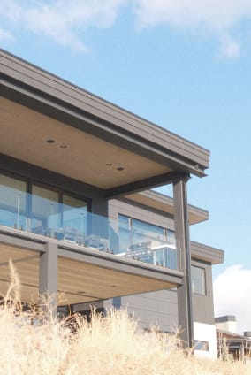 Frameless glass railing lines a covered second-story balcony seen from tall hillside grasses.