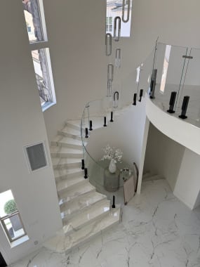 Curved glass railing on black talon clamps follows a white marble spiral stair under a crystal pendant.