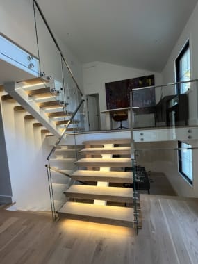 Light oak floating treads with under-tread LED lighting on a white mono stringer, flanked by pin-mounted glass and a stainless handrail.