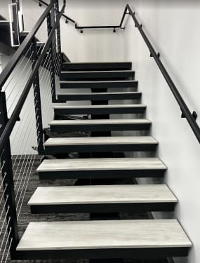 Gray treads on a black mono stringer between black cable railing and a wall-mounted black handrail in an office stairwell.