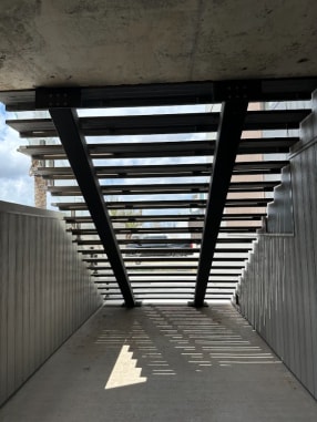 Looking up beneath a wide exterior steel stair with twin black stringers and open treads casting striped shadows.