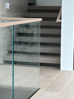 Gray-washed stack stair behind a frameless glass panel with a light wood cap rail.
