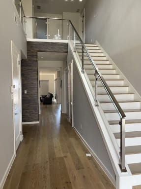 Stainless posts with horizontal rod infill and a matching handrail run up a white straight stair beside a long wood-floored hallway.