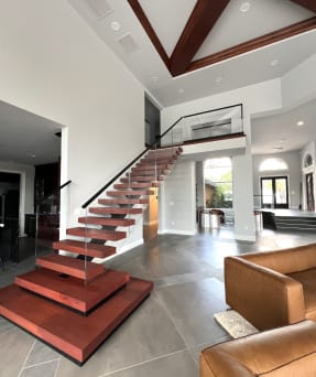 Mono stringer floating stair with red-stained treads and plinths, frameless glass and black handrail rising to a loft in an open great room.
