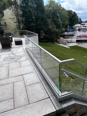 Glass base rail railing wraps a granite patio with outdoor seating, looking toward boats on a lake