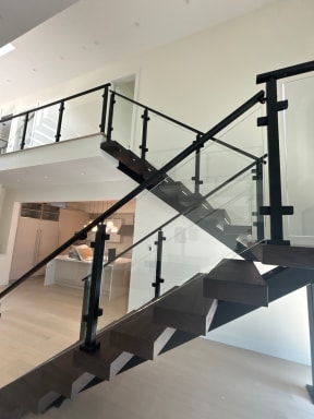 Black dual-stringer stair with dark treads and post-to-post glass railing rising to a loft