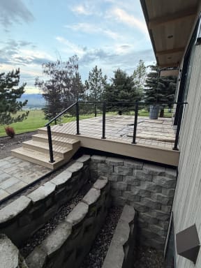 Black cable railing on a composite deck and steps above a stone window well, with mountain pasture beyond