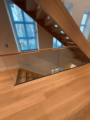 Frameless glass and oak handrail wrap a stair opening below a blue-taped arched window