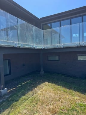 Frameless glass on side-mount brackets wraps an elevated deck on a gray modern home above a lawn