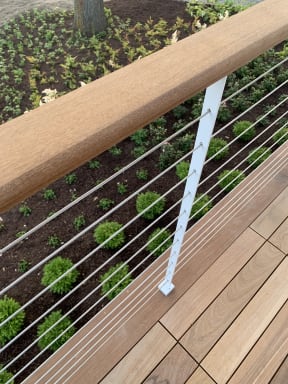 Close-up of a white cable railing post and wood cap rail along a hardwood deck edge above landscaping.