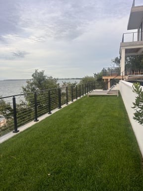 Black cable railing runs along a cliff-top lawn above the water beside a modern home.