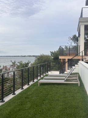 Black cable railing lines a lawn terrace with loungers, stepping up to a cable-railed deck beside the bay.