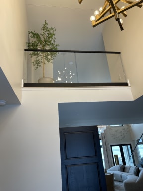 A frameless glass balcony railing with a slim black cap overlooks a two-story foyer with a chandelier.