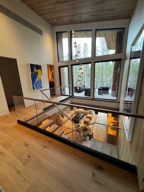A frameless glass guard with wood cap overlooks a double-height living room with floor-to-ceiling windows.