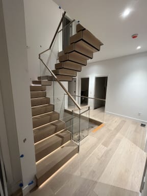 A U-shaped stack stair of thick white oak treads with LED step lighting rises between frameless glass guards with a wood handrail.